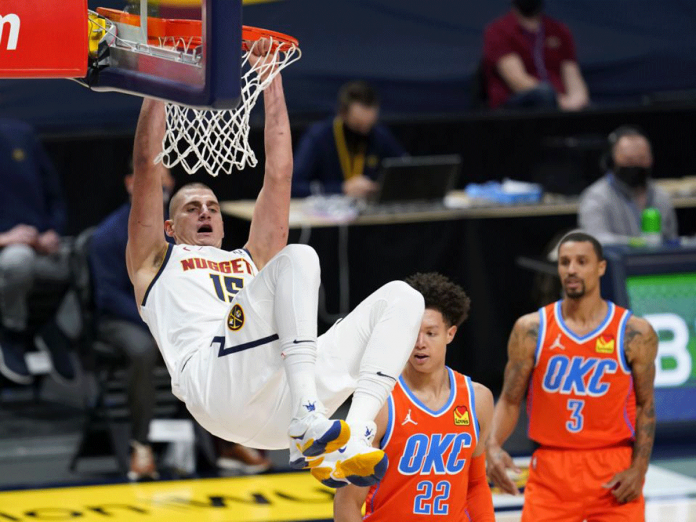 Denver Nuggets center Nikola Jokić hangs from the rim after dunking the ball for a basket against Oklahoma City Thunder forward Isaiah Roby and guard George Hill in the first half of an NBA basketball game on Jan. 19, 2021. David Zalubowski/AP Photo