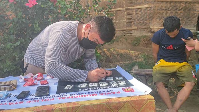 Drug suspect Raymond Tatel (right) of Barangay Tigum, Pavia, Iloilo appears to be sulking as a police operative conducts an inventory of the items seized in a buy-bust operation that led to the suspect’s arrest. PN PHOTO