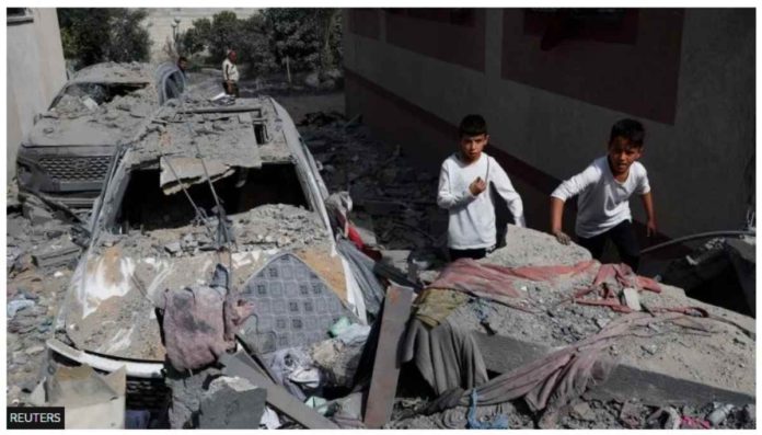 Two boys are seen standing next to a car that is heavily damaged and covered in debris in Gaza. In the foreground is what appears to be the rubble of a collapsed building. REUTERS