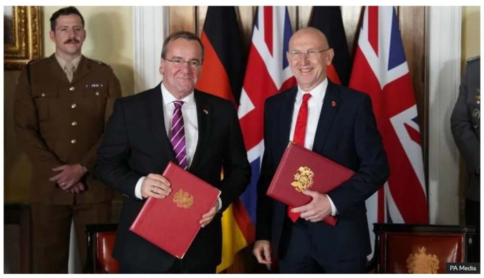 United Kingdom’s Defence Secretary John Healey and Germany’s Defence Minister Boris Pistorius smile, standing in front of the Union Jack and German flags, holding red binders. BBC