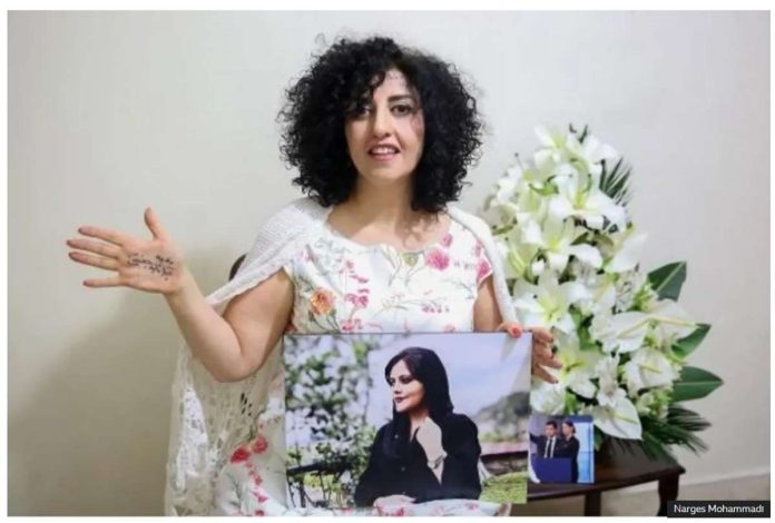 Narges Mohammadi Narges Mohammadi, wearing a white dress with flower patterns on it, holds out her right hand to show a message written on it, while holding a photo in her left hand. The photo is Mahsa Amini, whose death in custody triggered protests in Iran. Narges Mohammadi Photo