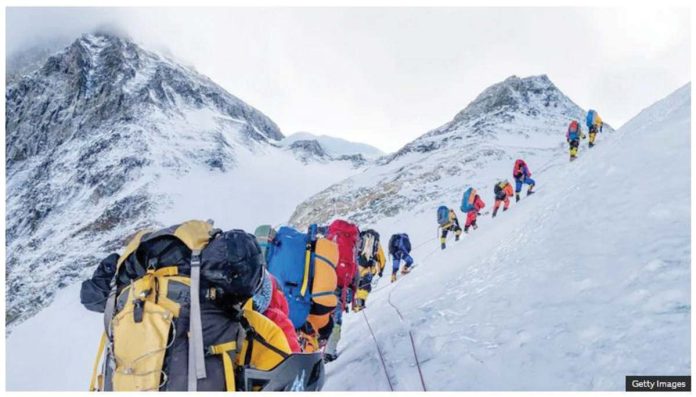 A line of mountain climbers hike across a snow-covered slope with mountain peaks in the background. Nepal is often criticized for allowing too many climbers on Everest. GETTY IMAGES