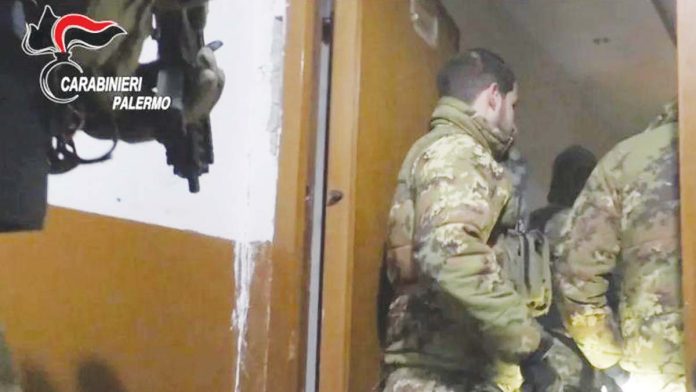 Two uniformed officers of the Carabinieri wearing jackets with a camouflage print stand in a doorway while a colleague behind them holds a gun. Italy's military police launched the operation in the early hours of Tuesday against the Mafia. CARABINIERI PALERMO