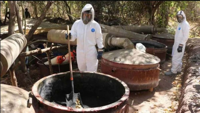 Two Mexican army personnel wearing white protective suits, full face visors and heavy-duty black gloves stand in front of large vats at a site which was used as a meth laboratory in the community of Higueras de Abuya in Mexico. REUTERS