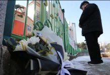 A man stands with his hands crossed in front of a school gate. Bouquets of flowers lay on the ground beside him. Locals laid flowers at the school’s gate to mourn an eight-year-old student. NEWS1