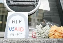 A sign placed reading “RIP USAID 1961-2925” and flowers are placed beneath the covered seal of the US Agency for International Development headquarters in Washington, DC. AFP